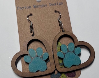 May include: A pair of wooden heart-shaped earrings with a blue paw print cutout. The earrings are hanging from silver hooks. The earrings are on a brown card with the text "Payton Murphy Design".