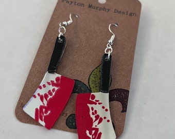 May include: A pair of silver earrings shaped like a bloody butcher knife. The earrings are on a brown card with the text "Payton Murphy Design".