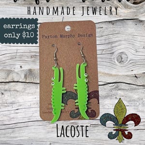 May include: Bright green alligator-shaped earrings with silver hooks. The earrings are displayed on a brown card with the text "Payton Murphy Design" and "LACOSTE". The background features the text "unique & unusual HANDMADE JEWELRY".