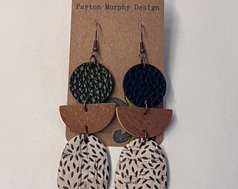 May include: A pair of dangle earrings with a black, green, and brown geometric design. The earrings feature a round black and green leather piece, a half-circle brown wood piece, and a white and black patterned cork piece.