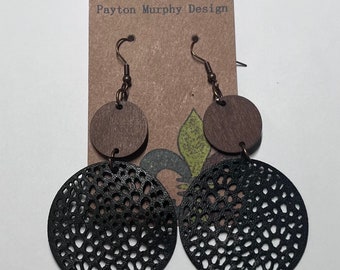 May include: A pair of black and brown wooden earrings with a delicate, intricate lace pattern. The earrings are hanging from a brown card with the text "Payton Murphy Design".