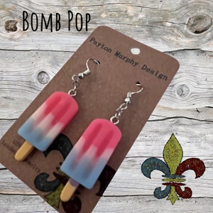 May include: A pair of earrings featuring a pink, white and blue popsicle design. The earrings are on a brown card with the text "Payton Murphy Design" printed on it. The background is a wooden surface.