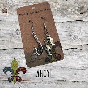 Könnte beinhalten: Silberfarbene Ohrringe mit einem Angelhaken und Fischskelett-Anhängern. Die Ohrringe werden auf einer braunen Karte mit dem Text "Payton Murphy Design" und "AHoy!" präsentiert. Der Hintergrund ist eine rustikale Holzoberfläche.