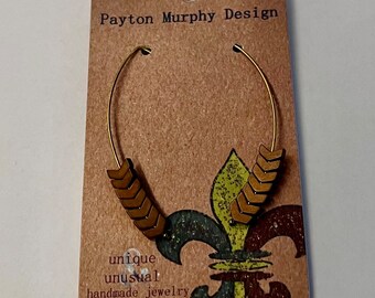 May include: A pair of gold hoop earrings with brown arrow-shaped charms. The earrings are displayed on a brown card with the text "Payton Murphy Design", "unique", "unusual", and "handmade jewelry".