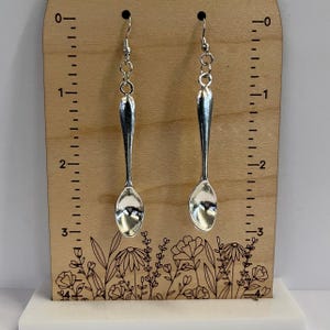 May include: A pair of silver spoon earrings hanging from a wooden display board with a ruler and floral design. The earrings are approximately 7.5 cm long.