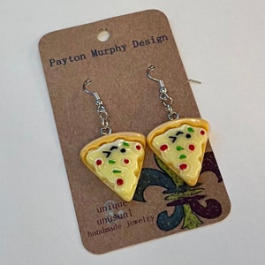 May include: A pair of silver dangle earrings with a yellow pizza slice charm. The pizza slice has red and green toppings and a white cheese base. The card behind the earrings says "Payton Murphy Design" and "unique unusual handmade jewellery".