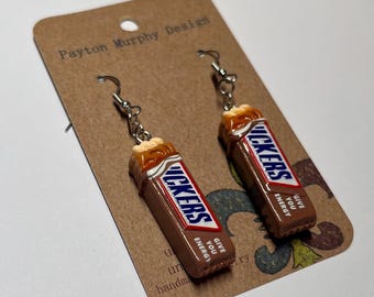 May include: A pair of novelty earrings shaped like miniature Snickers candy bars. The earrings feature a brown, white, and red color scheme, with the word "Snickers" prominently displayed. The text "GIVE YOU ENERGY" is also visible. The earrings are attached to silver-colored fish hook ear wires.