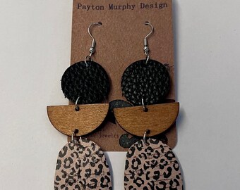May include: A pair of dangle earrings with a black leather circle, a brown wood half-circle, and a pink and black leopard print design. The earrings are labeled "Payton Murphy Design".
