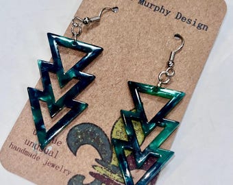 May include: A pair of dangle earrings with a geometric design. Each earring features three connected, open-frame triangles in shades of green and blue. The earrings are suspended from silver-colored fish hook ear wires. The earrings are displayed on a brown card with the text "Payton Murphy Design".