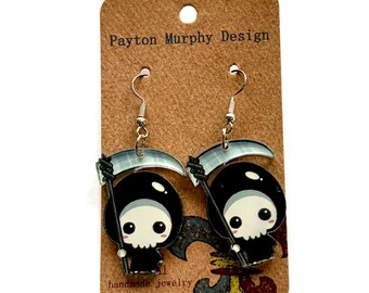 May include: A pair of dangle earrings featuring a cartoon grim reaper design. The earrings are black, white, and gray, with silver-colored hooks. The earrings are attached to a brown card with the text "Payton Murphy Design" and "handmade jewelry".