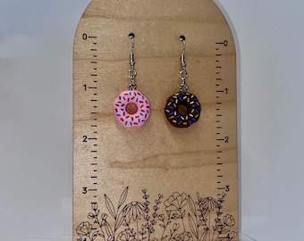 May include: A pair of novelty earrings shaped like miniature donuts. One is pink with colorful sprinkles, and the other is chocolate with sprinkles. The earrings hang from silver-colored hooks and are displayed on a wooden stand with a floral design.