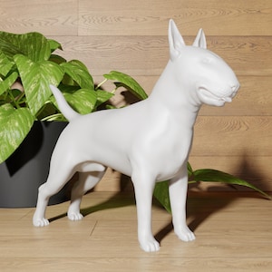Op de afbeelding: Witte keramische sculptuur van een Bullterriër die op een houten oppervlak staat. De hond kijkt naar rechts en heeft zijn tong lichtjes uitgestoken.