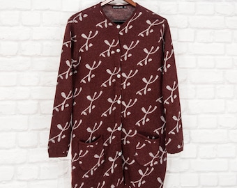 May include: A burgundy cardigan with a white abstract pattern and two front pockets hangs on a wooden hanger. The long-sleeved sweater has a button-up front and a round neckline. The background is a white brick wall.