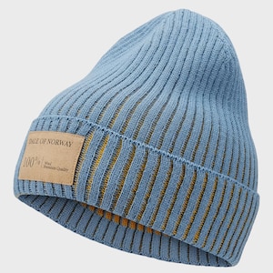 Puede incluir: Un gorro de punto acanalado azul claro con una etiqueta de cuero marrón. La etiqueta dice "DALE OF NORWAY" y "100% Wool Premium Quality". El gorro tiene un forro amarillo-naranja y es un accesorio de invierno.