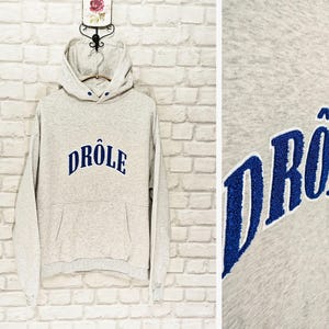 May include: A light gray hoodie with a drawstring hood and a front pocket. The word "DRÔLE" is embroidered in blue across the chest. The hoodie is hanging on a hanger against a white brick wall.
