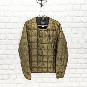 May include: Olive green quilted jacket with a button-up front and a small logo on the chest. The jacket is hanging on a hanger against a white brick wall. The jacket appears to be lightweight and suitable for layering.