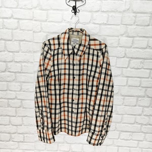 Può includere: Una camicia a maniche lunghe con un motivo a quadri color crema, nero e arancione. La camicia ha un colletto e polsini abbottonati. La camicia è appesa a un gancio di metallo contro un muro di mattoni bianchi.