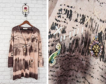 Peut inclure: Un cardigan à manches longues avec un motif tie-dye dans les tons de brun et de rose. Le cardigan a une fermeture boutonnée et deux poches. Il est orné de coutures décoratives et d'un petit charme en métal.