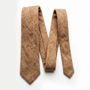 May include: A brown tie with a textured, woven appearance. The tie is laid out on a white surface, showcasing its length and design. The tie's color is a warm, earthy tone, suitable for various outfits.