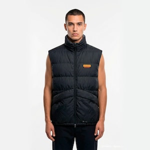 May include: Black puffer vest with a high collar and a small rectangular patch on the chest. The vest is sleeveless and has a full-length zipper. The vest is worn over a dark shirt.