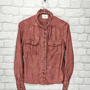 May include: A long-sleeved, rust-colored corduroy shirt with a ruffled collar and gold buttons. The shirt has two flap pockets on the front and a small label that reads "SEZANE" on the collar.