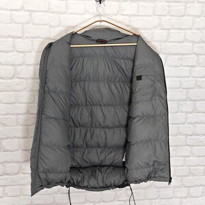 May include: A gray puffer jacket hanging on a wooden hanger against a white brick wall. The jacket is open, revealing the quilted interior. Black drawstrings are visible at the bottom. The jacket is designed for warmth and protection from the elements.