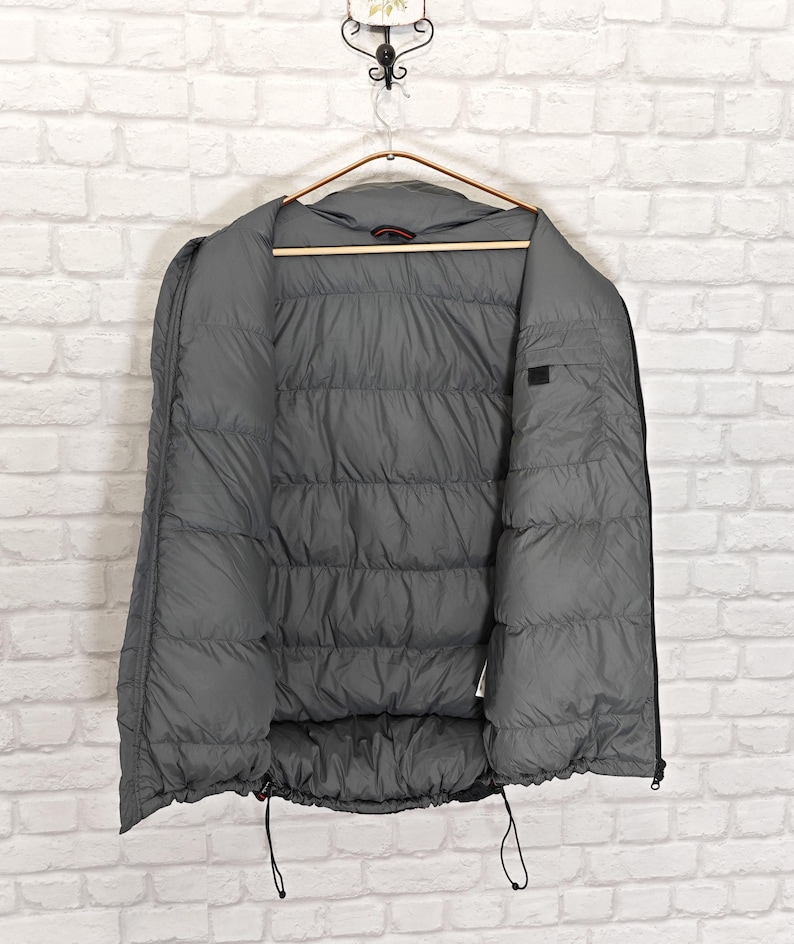 May include: A gray puffer jacket hanging on a wooden hanger against a white brick wall. The jacket is open, revealing the quilted interior. Black drawstrings are visible at the bottom. The jacket is designed for warmth and protection from the elements.