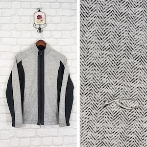 May include: A gray and black zip-up jacket with a herringbone pattern on the sleeves. The jacket has a stand-up collar and a black zipper. The jacket is hanging on a hanger against a white brick wall.