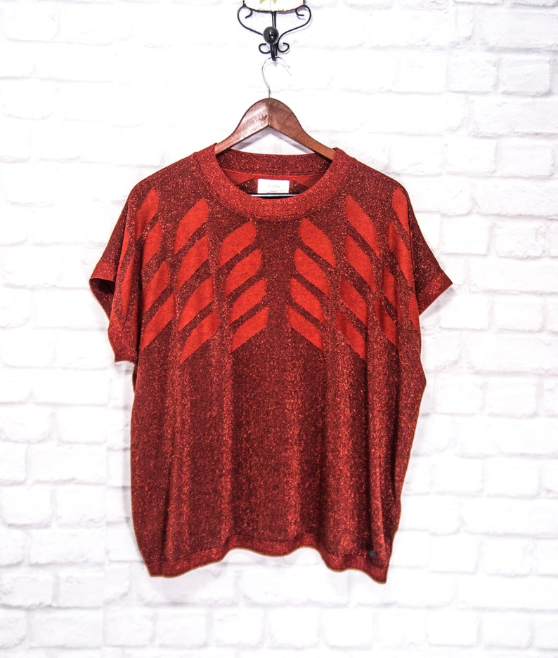 May include: A red sparkly short-sleeved sweater with a geometric pattern. The sweater has a round neckline and is made of a soft, comfortable material.