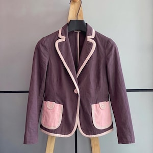 May include: A dark purple blazer featuring light pink piping along the lapels, hem, and two front patch pockets. The blazer has long sleeves, a single button closure at the center, and a notched collar design with a structured, tailored fit.