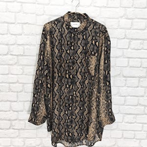 Può includere: Camicia a maniche lunghe con stampa a pelle di serpente nei toni del nero, marrone e beige. La camicia è appesa a una gruccia di metallo contro un muro di mattoni bianchi. Colletto e chiusura con bottoni.