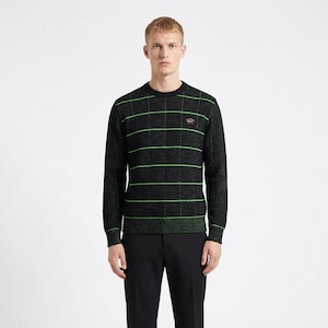 Puede incluir: Suéter negro con rayas horizontales y verticales de color verde neón. El suéter tiene cuello redondo y mangas largas. Un pequeño emblema es visible en el pecho. La persona de la imagen lleva pantalones negros.