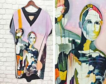 May include: A lavender t-shirt with a v-neck and short sleeves. The shirt features a colorful abstract painting of three figures with black outlines.