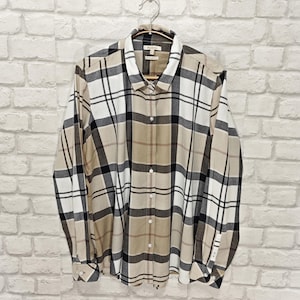 Puede incluir: Camisa de manga larga con estampado de cuadros en beige, negro y blanco. La camisa está colgada de una percha de madera sobre una pared de ladrillos blancos. Tiene cuello clásico y puños abotonados.