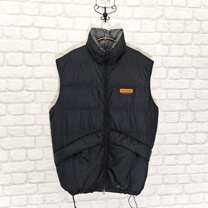 May include: Black puffer vest with a high collar and a full-length zipper. The vest has two front pockets and a small rectangular patch with text. The vest is hanging on a metal hanger against a white brick wall.