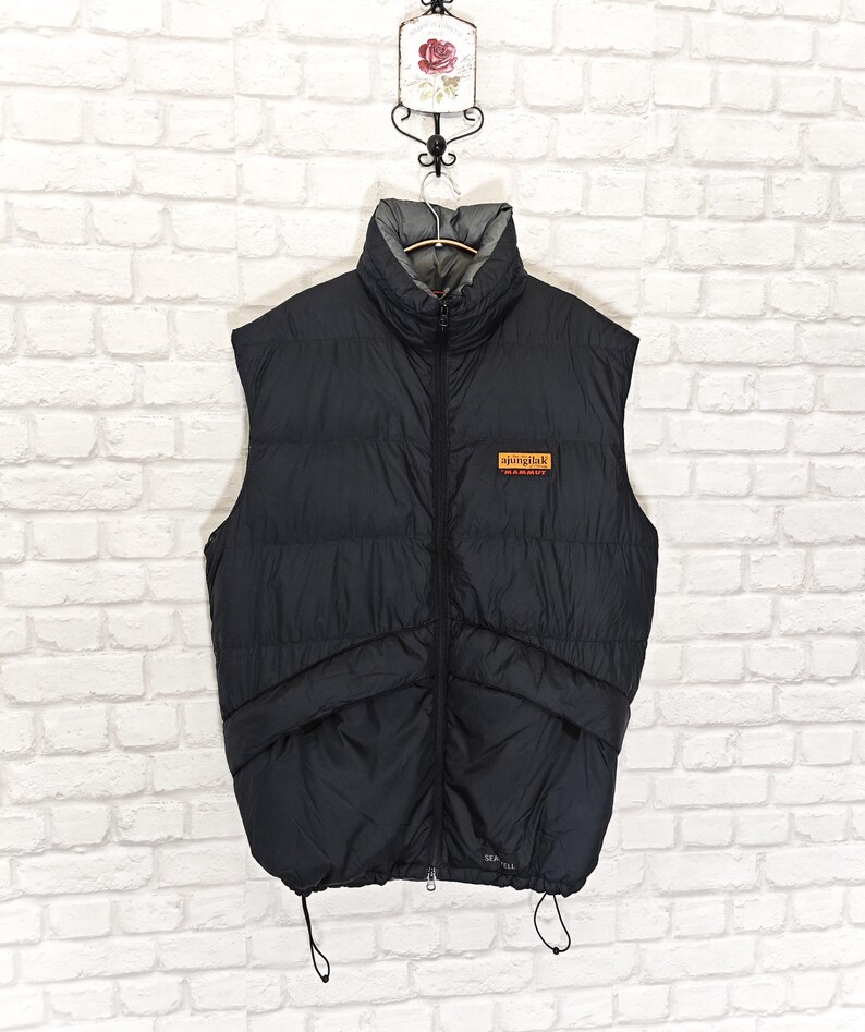 May include: Black puffer vest with a high collar and a full-length zipper. The vest has two front pockets and a small rectangular patch with text. The vest is hanging on a metal hanger against a white brick wall.