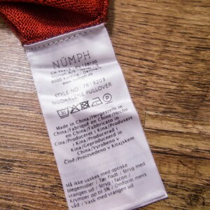 May include: White care label with black text for a N&Uuml;MPH NUDARLENE PULLOVER. The label includes the style number 7619203 and instructions for washing and caring for the garment. The label is made in China.
