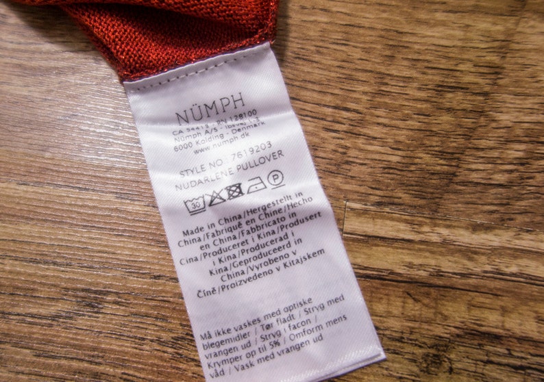 May include: White care label with black text for a N&Uuml;MPH NUDARLENE PULLOVER. The label includes the style number 7619203 and instructions for washing and caring for the garment. The label is made in China.