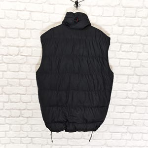 May include: Black puffer vest with a high collar and a quilted design. The vest has a full-length zipper and two drawstrings at the bottom. The vest is hanging against a white brick wall.