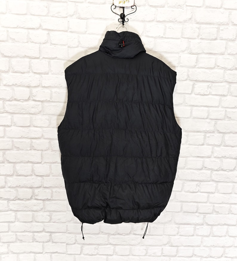 May include: Black puffer vest with a high collar and a quilted design. The vest has a full-length zipper and two drawstrings at the bottom. The vest is hanging against a white brick wall.