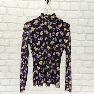 May include: A black, sheer turtleneck top with long sleeves, patterned with purple and yellow flowers. The top is suspended on a metal hanger. The background is a white brick wall, highlighting the floral design of the garment.