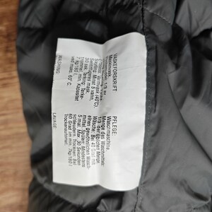 May include: A close-up of a dark gray garment label with text in multiple languages. The label provides washing instructions, including machine wash at 40 degrees Celsius, and tumble dry information.