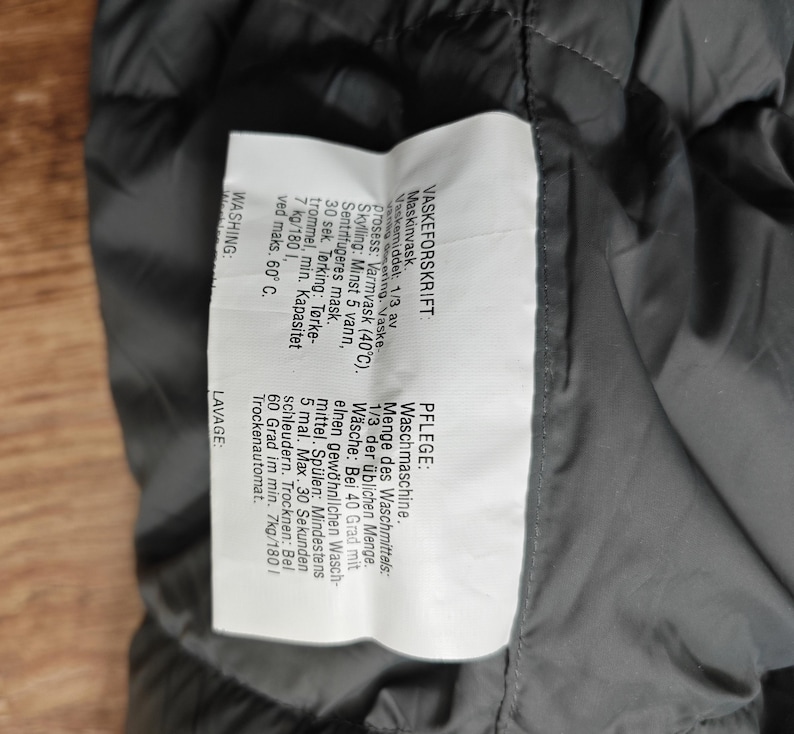 May include: A close-up of a dark gray garment label with text in multiple languages. The label provides washing instructions, including machine wash at 40 degrees Celsius, and tumble dry information.