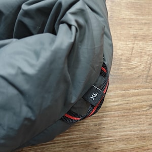 May include: A folded, gray sleeping bag with a black and red strap. The strap has the letters "XL" printed on it. The sleeping bag is on a wooden surface.