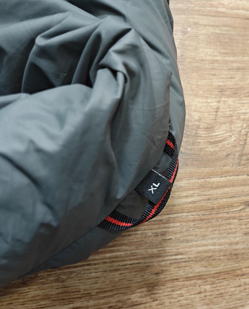 May include: A folded, gray sleeping bag with a black and red strap. The strap has the letters "XL" printed on it. The sleeping bag is on a wooden surface.