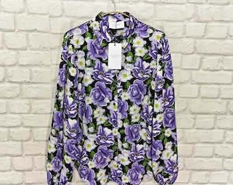 May include: A long-sleeved button-down shirt with a floral pattern. The shirt features a black background with large purple roses and smaller white flowers. The shirt is hanging on a hanger against a white brick wall.