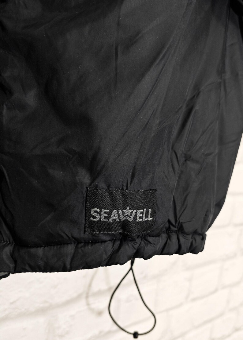 May include: Black jacket with a drawstring hem. The jacket features a small rectangular patch with the text "SEAWELL" and a star graphic. The fabric appears to be a lightweight, possibly water-resistant material.