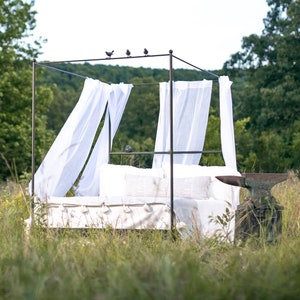 A black canopy bed frame with white sheer curtains in a grassy field. The bed frame is made of metal and has a simple design. The bed is made up with white linens and pillows. The bed is surrounded by tall green grass and trees in the background.
