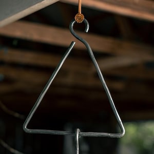 May include: A black metal triangle hanging from a wooden beam. The triangle is suspended by a brown leather strap with a metal hook. The triangle is a musical instrument used to create a ringing sound.