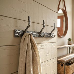May include: A wall-mounted coat rack featuring a dark, hammered metal finish with three curved hooks. The rectangular base plate is textured and secured with visible screws. A beige, woven fabric scarf hangs from the first hook.